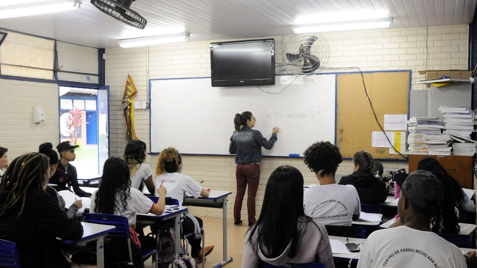 Professora escreve no quadro branco diante de estudantes sentados em sala de aula de escola pública