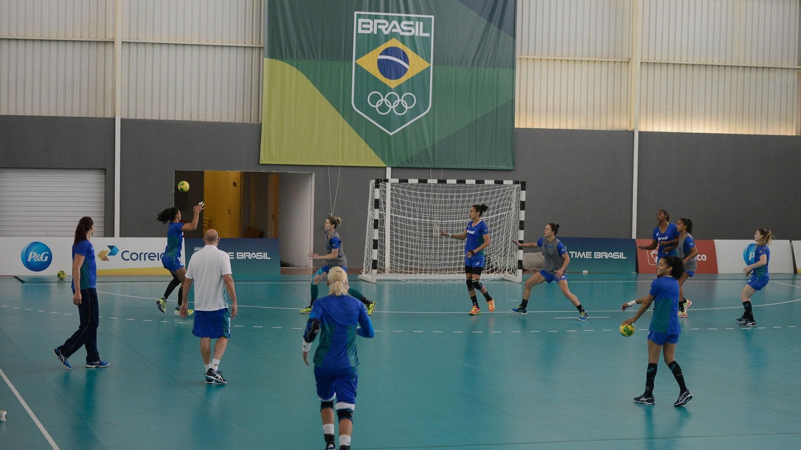 Jogadoras de handebol de uniforme azul treinam em quadra coberta sob faixa do Time Brasil