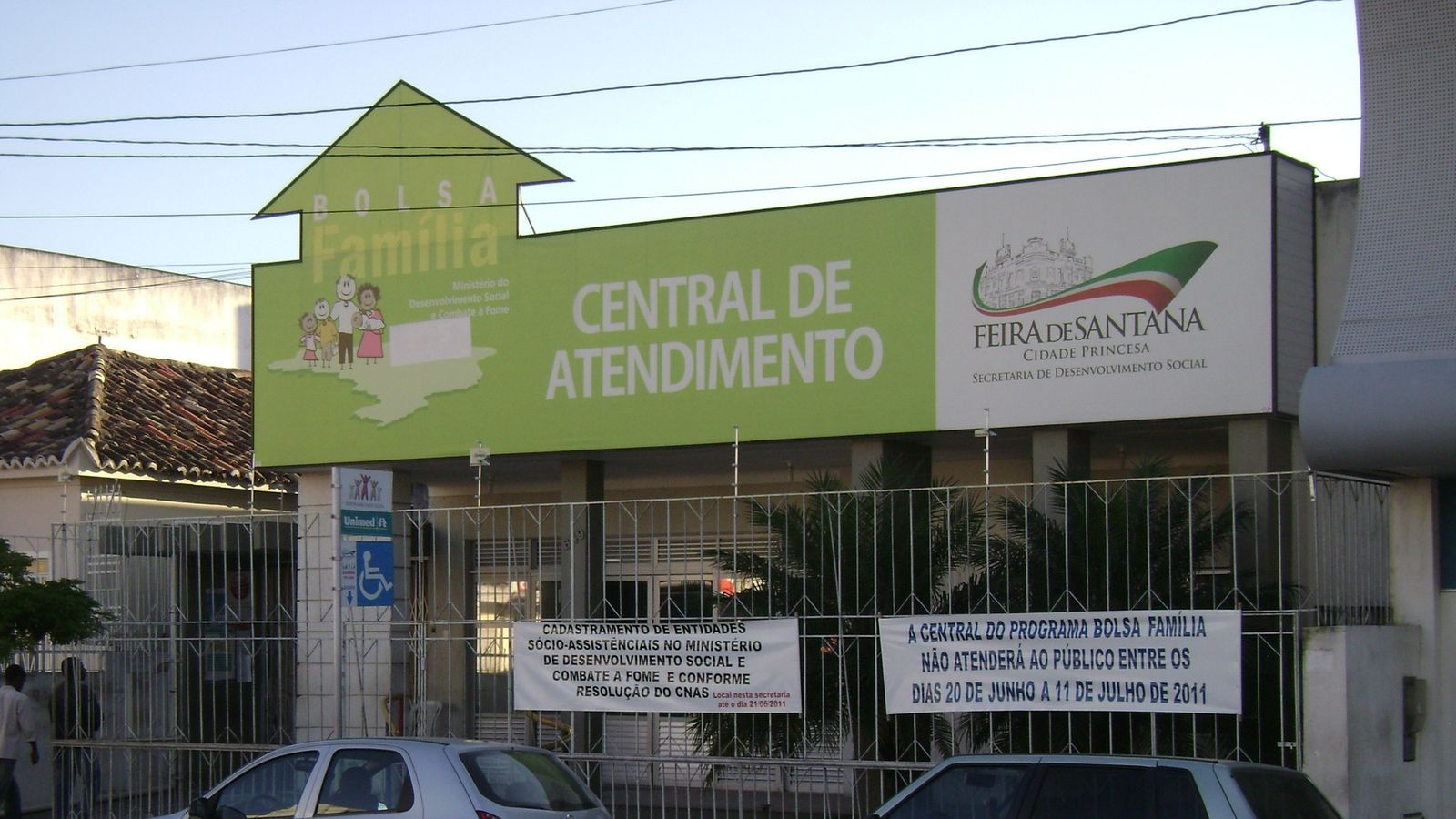 Fachada da Central de Atendimento do Bolsa Família em Feira de Santana, com letreiro verde e carros em frente